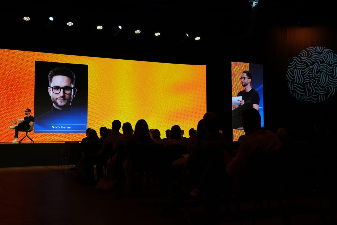 Niko Hems on stage at Life Summit, speaking to a seated audience with his name on the screen behind him